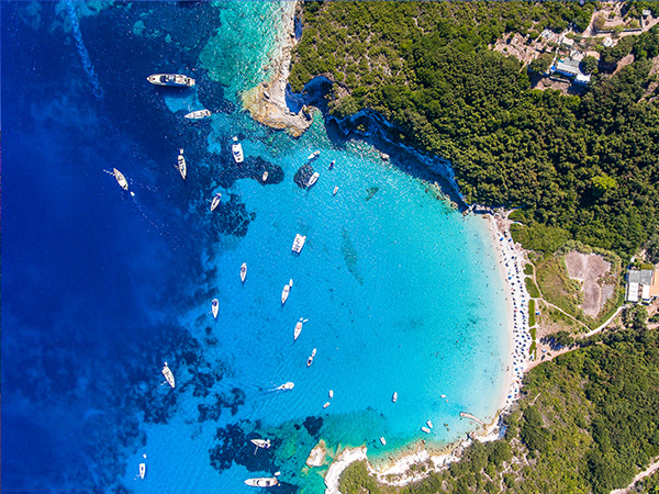 Antipaxos Island, Greece, with sandy beach, people swimming and yachts docked in the ethereal clear blue waters of the Ioanian island near Corfu