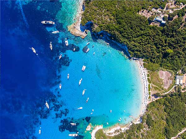 Antipaxos Island, Greece, with sandy beach, people swimming and yachts docked in the ethereal clear blue waters of the Ioanian island near Corfu