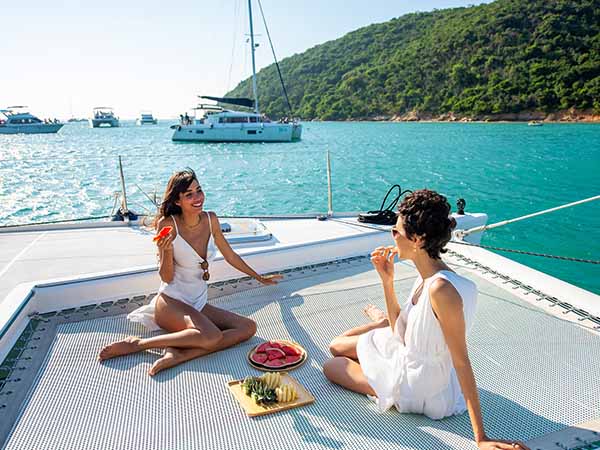Caucasian woman friends enjoy luxury lifestyle catamaran boat sailing with eating fresh fruit together at summer sunset