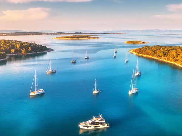 Aerial view of beautiful yachts and boats on the sea at sunset in summer. Adriatic sea, Kamenjak, Croatia. Top view of luxury yachts, sailboats, lagoon, clear blue water, and green forest. Travel