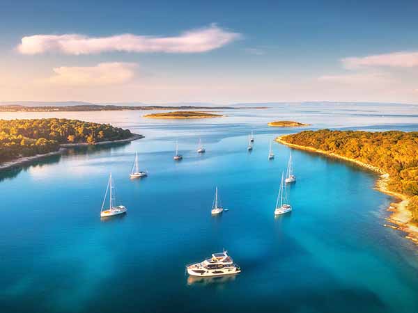 Aerial view of beautiful yachts and boats on the sea at sunset in summer. Adriatic sea, Kamenjak, Croatia. Top view of luxury yachts, sailboats, lagoon, clear blue water, and green forest. Travel