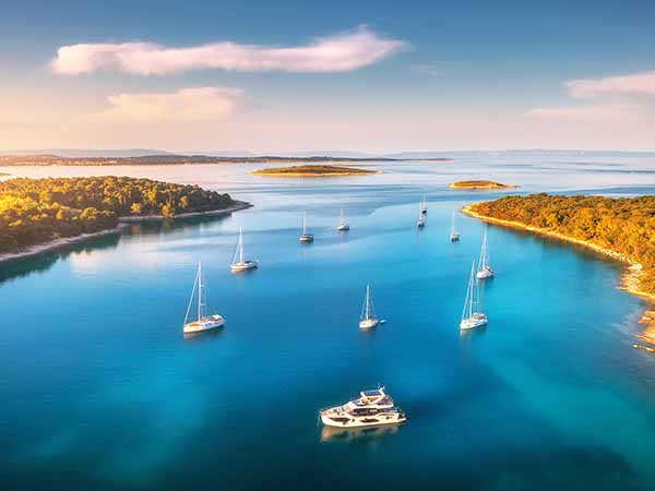 Aerial view of beautiful yachts and boats on the sea at sunset in summer. Adriatic sea, Kamenjak, Croatia. Top view of luxury yachts, sailboats, lagoon, clear blue water, and green forest. Travel